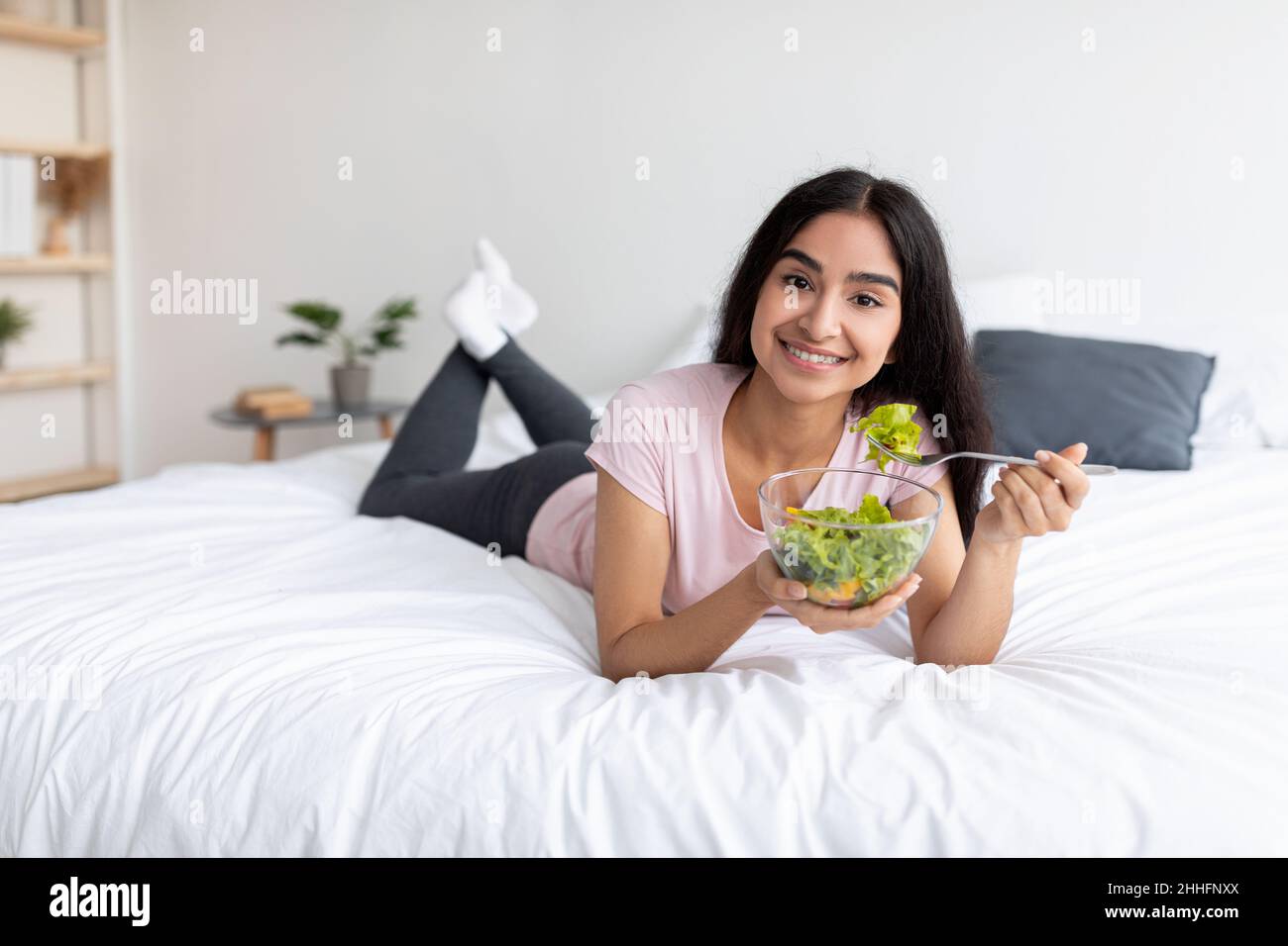 Belle jeune femme indienne mangeant de la salade de légumes tout en étant couché sur le lit à la maison, espace libre.Régime alimentaire brut équilibré Banque D'Images