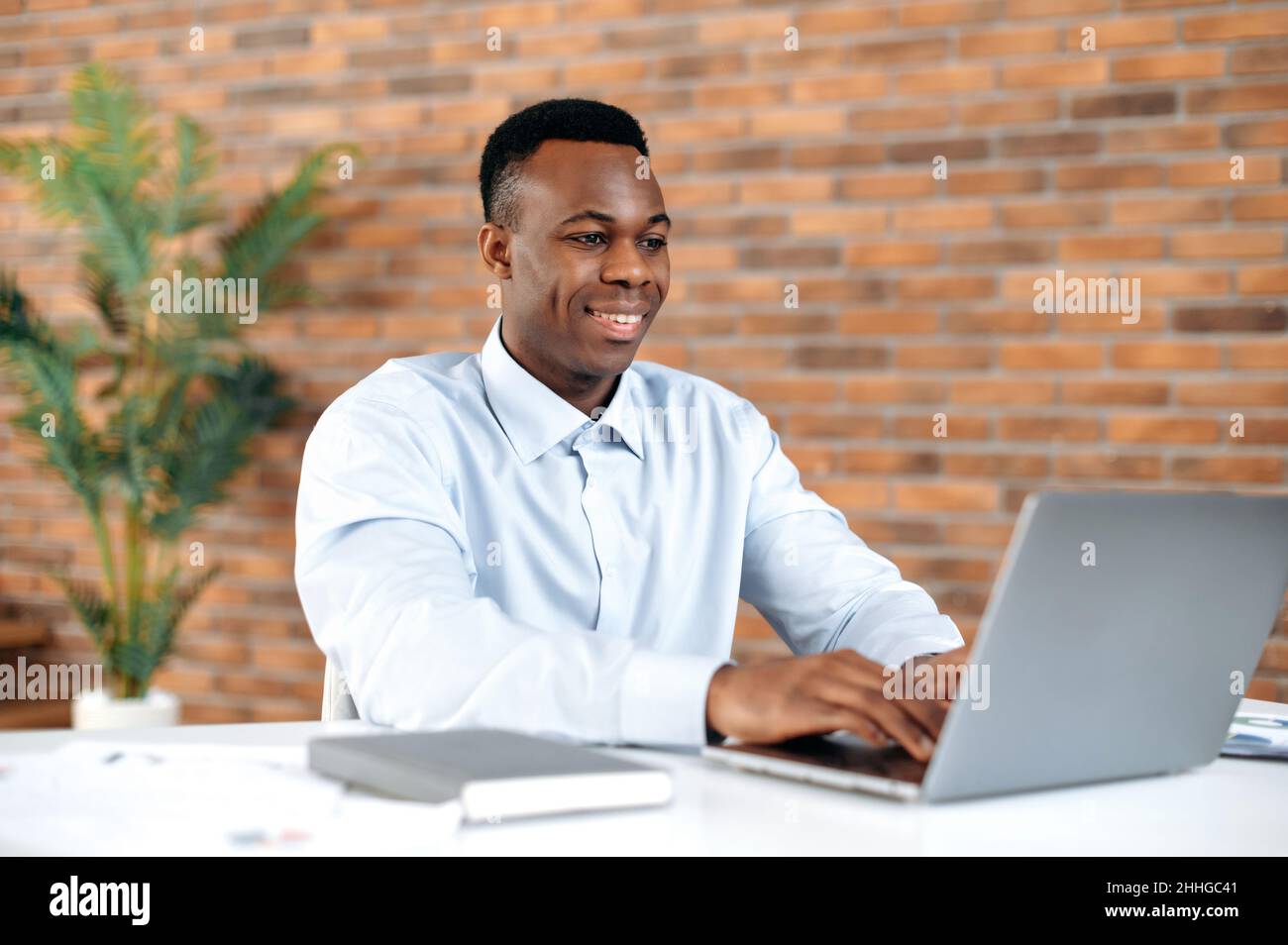 Homme noir confiant et positif utilisant un ordinateur portable dans un bureau moderne.Homme d'affaires, indépendant, Manager, en chemise formelle, en parcourant les e-mails,travailler et taper sur un ordinateur portable, travailler sur un projet, sourire Banque D'Images