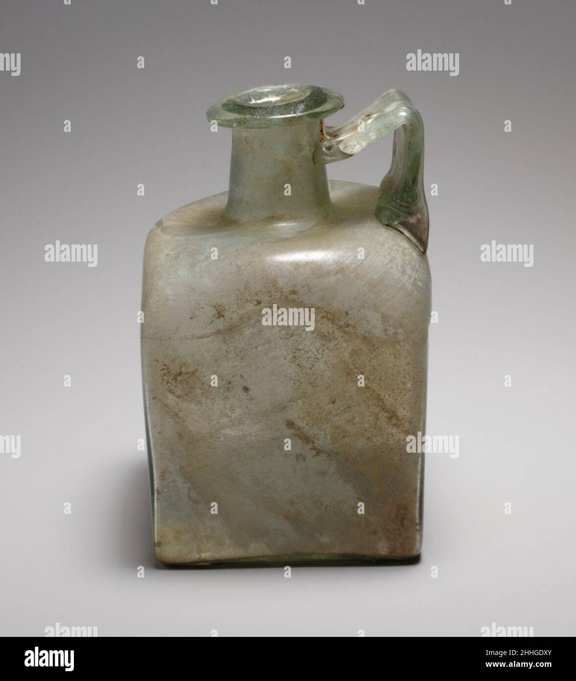 Flacon carré en verre fin 1st–3rd Century A.D. Roman translucide vert pâle, avec manche de même couleur.rebord replié, au-dessus et à l'intérieur, formant légèrement une constriction à la bouche, avec surface supérieure biseautée; col cylindrique, avec repères d'outillage autour de la base; épaulement enfoncé avec coins arrondis et inclinés; côtés plats et verticaux; fond plat;Large poignée à lanière, avec décoration peignée sur la surface extérieure, fixée au bord extérieur de l'épaule, dressée verticalement, tournée en angle aigu et tractée sur le cou, avec une traînée se terminant sur le dessous de la jante. Décoration moulée sur le fond comprenant quatre angles en forme de L au niveau du maïs Banque D'Images
