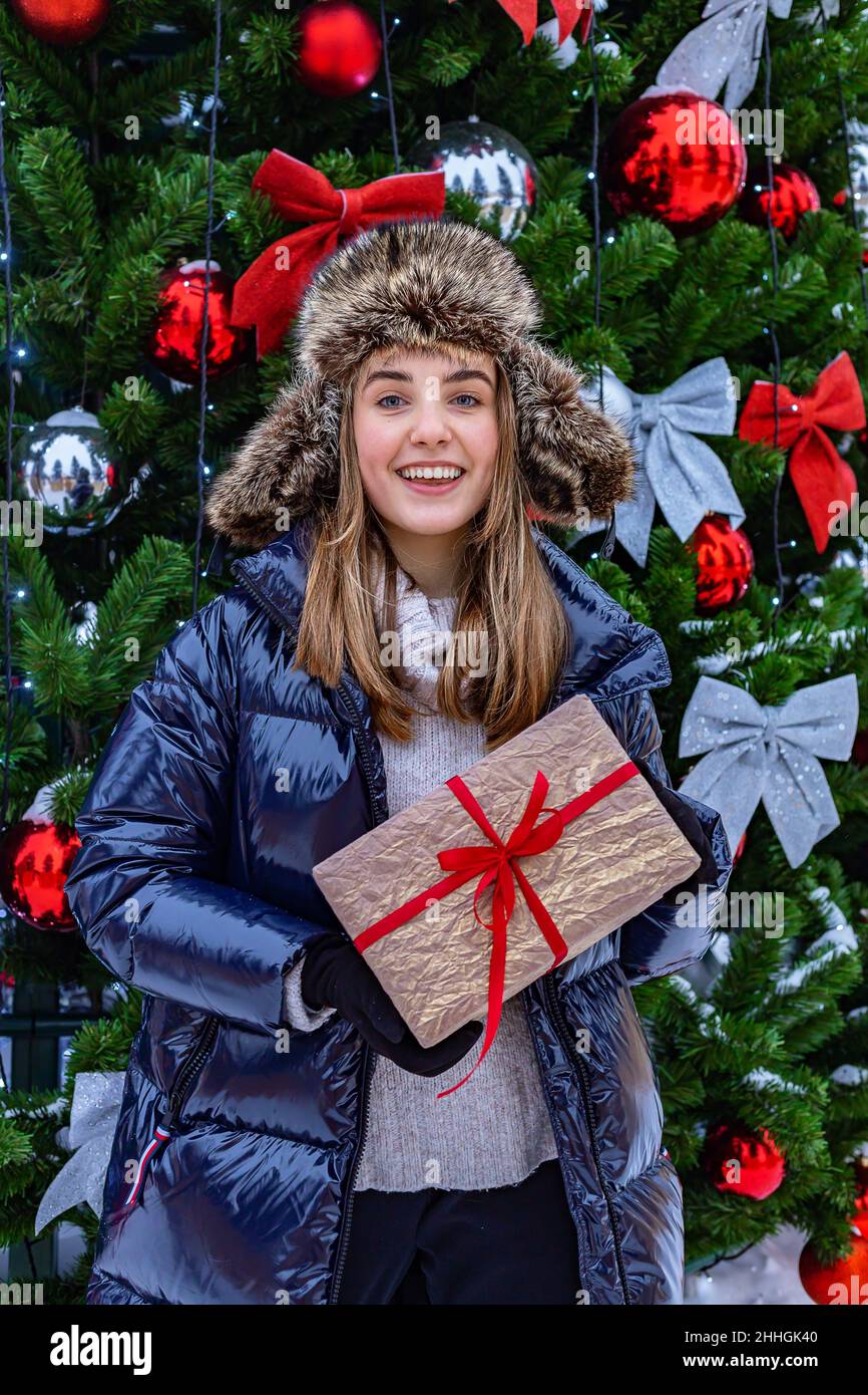 Bonne fille blonde souriante avec cadeau de Noël.Jolie fille adolescente d'origine caucasienne dans un chapeau de fourrure et une veste en duvet tient un Noël Banque D'Images