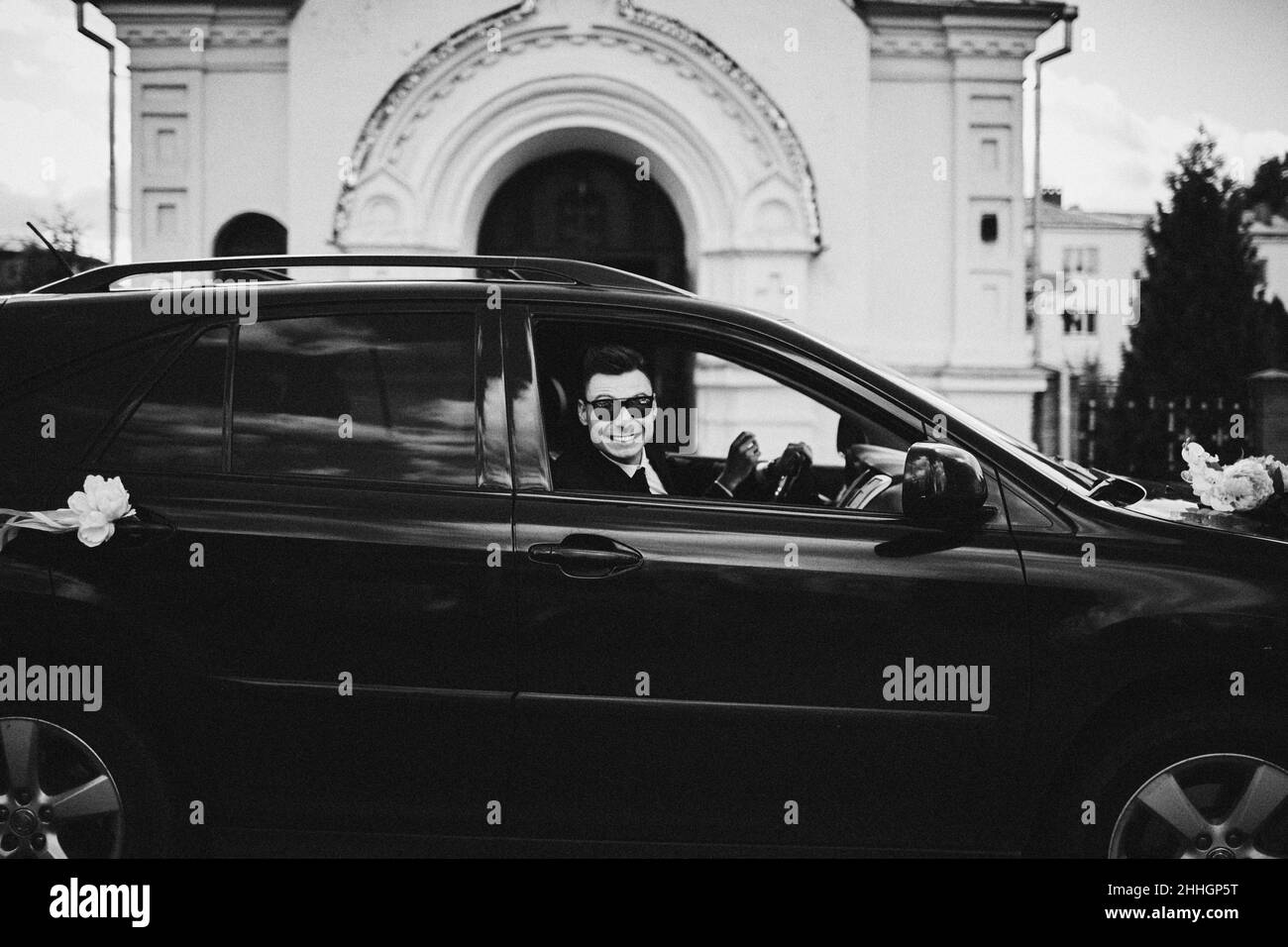 beau marié dans des lunettes de soleil est assis dans une voiture de luxe décorée. homme souriant en costume est en voiture le jour du mariage. photo noir et blanc Banque D'Images
