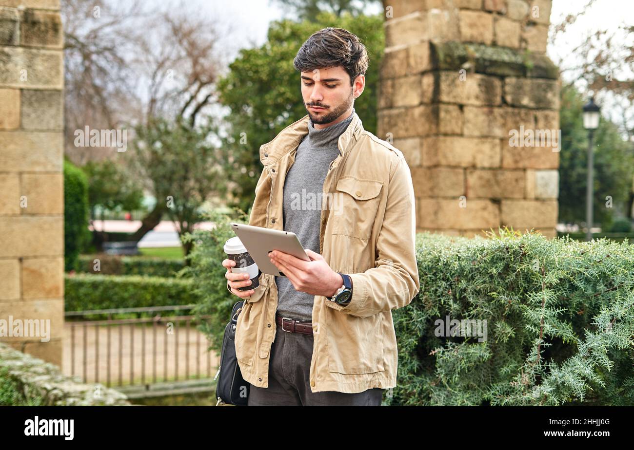 Homme sérieux dans des vêtements formels debout avec un sac pour ordinateur portable et une tasse de boisson à emporter dans la main dans le parc et la tablette de navigation Banque D'Images