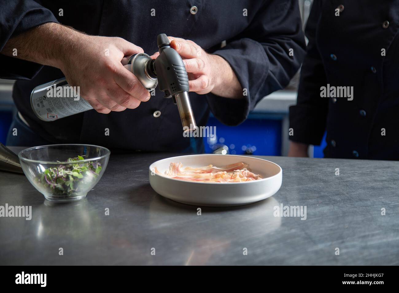 Homme méconnaissable utilisant une torche à gaz pour cuire des tranches de porc dans un bol tout en travaillant dans la cuisine du restaurant avec un collègue Banque D'Images