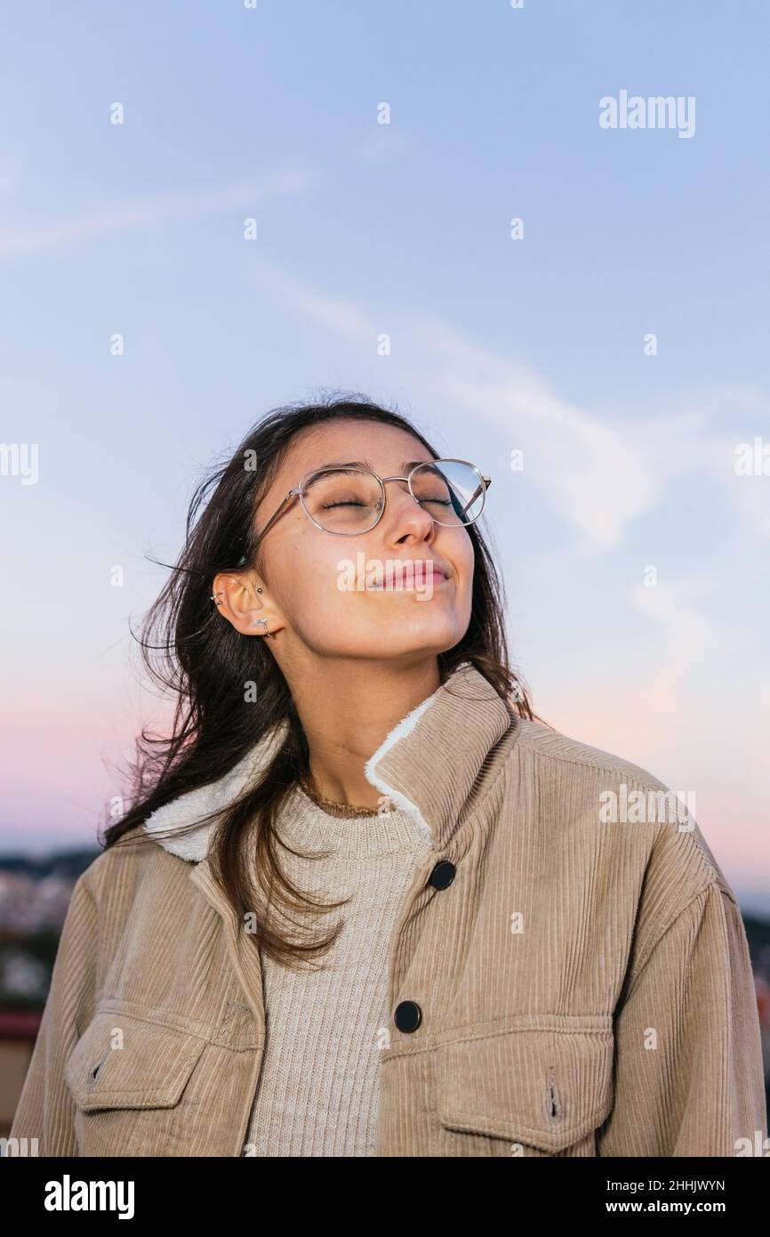 Bonne adolescente en lunettes souriant largement avec les yeux fermés contre le ciel le soir Banque D'Images