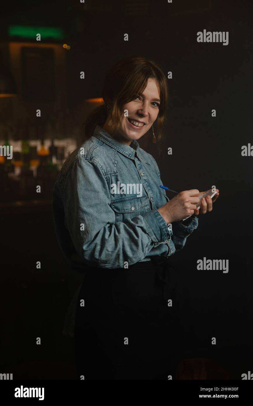 Bonne serveuse regardant l'appareil photo et prenant la commande en écrivant dans le carnet près du comptoir du bar pendant le travail dans un café moderne Banque D'Images
