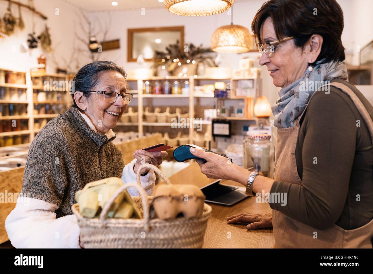Vue latérale d'une femme positive qui se tient à portée de main avec le terminal pour une femme âgée heureuse qui fait le paiement avec une carte de crédit dans un magasin écologique Banque D'Images