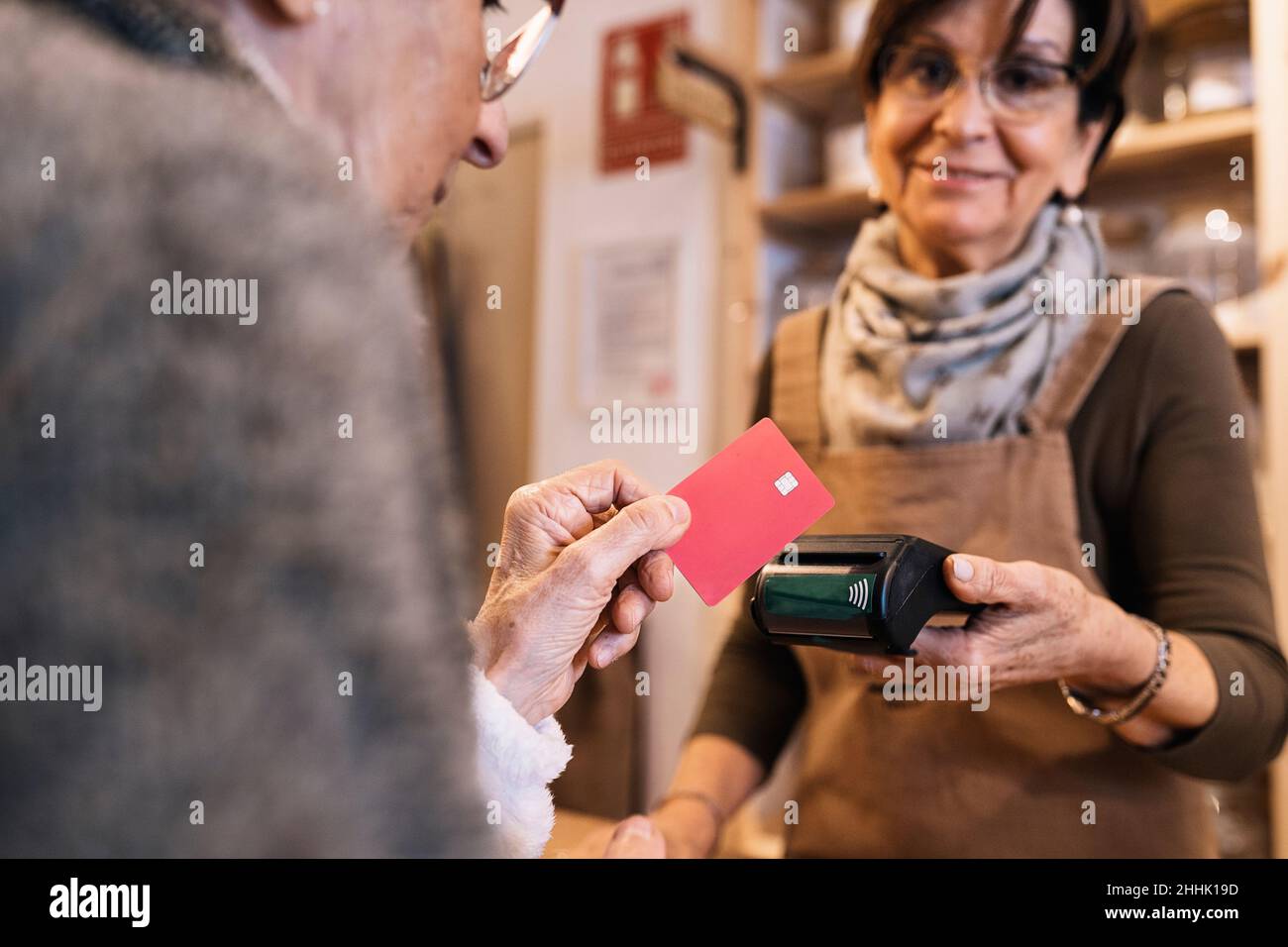 Vue latérale de la courte femelle positive unreconnaissable atteignant la main avec le terminal à la femme heureuse senior faisant le paiement avec la carte de crédit dans l'ECO Banque D'Images
