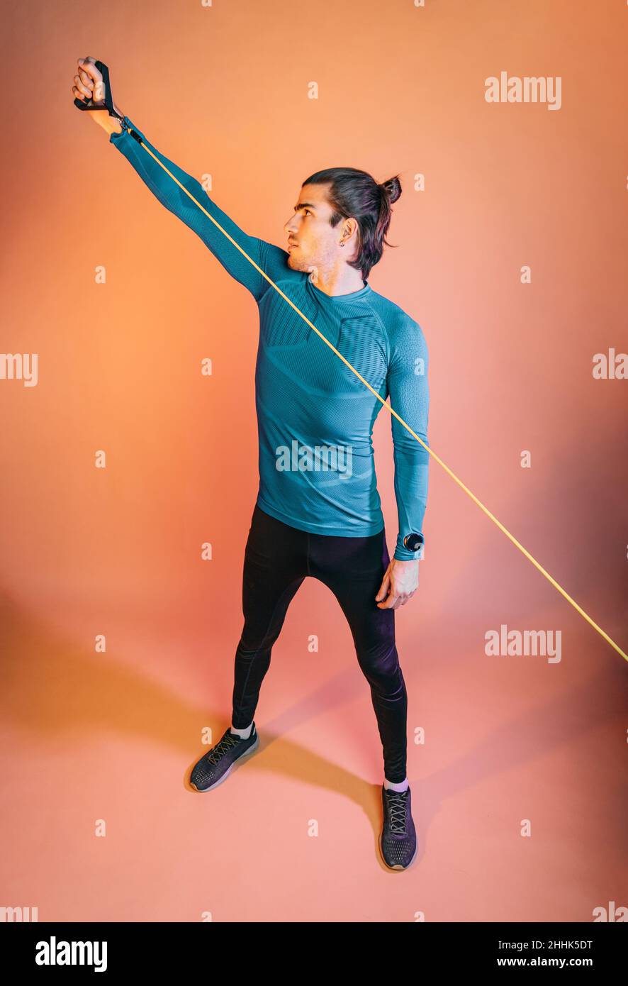 Pleine longueur de jeune tenue jeune hispanique athlète masculin avec des cheveux foncés dans les vêtements de sport exercice avec corde de résistance debout contre fond orange i Banque D'Images