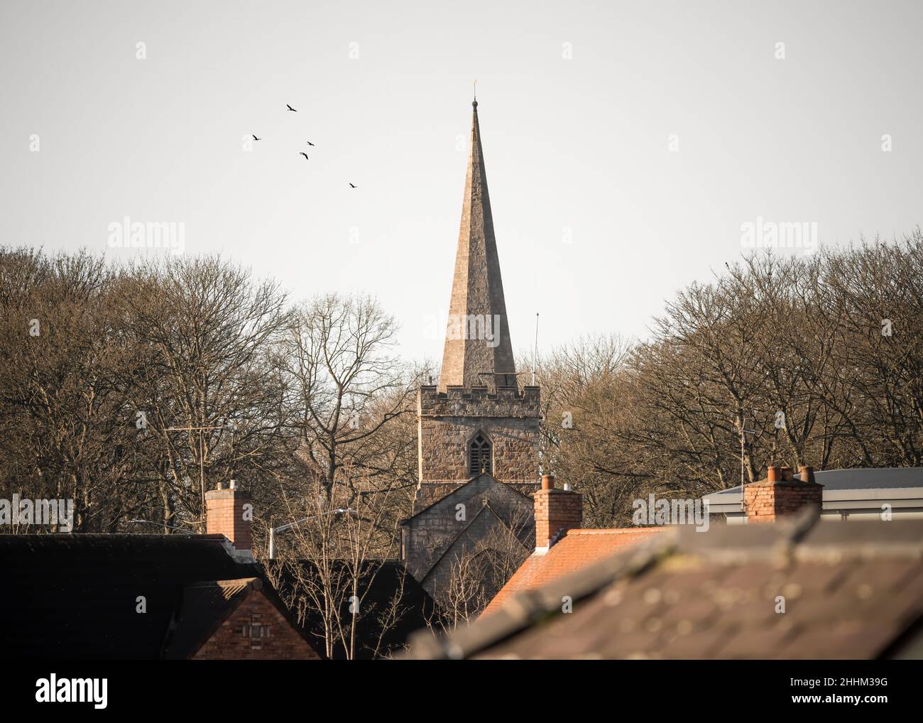 La pierre de l'église pointue flèche dans le centre du village s'élevant au-dessus des maisons résidentielles de la ville urbaine.Coucher de soleil et oiseaux dans le ciel. Banque D'Images
