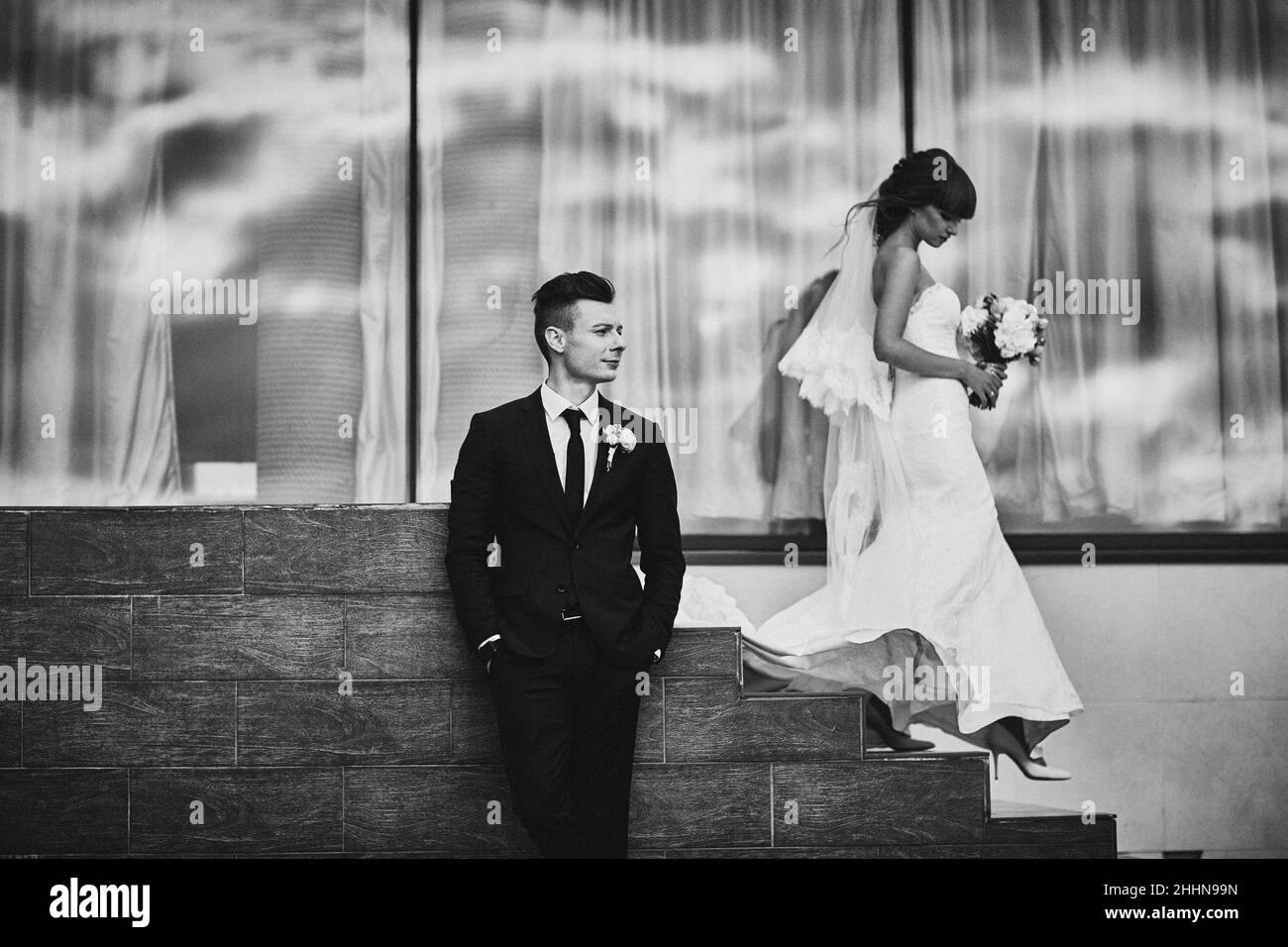 Jeune couple en amour mariée et marié posant à l'extérieur près de la grande fenêtre panoramique. Femme en robe longue blanche et bouquet de fleurs, homme en noir Banque D'Images