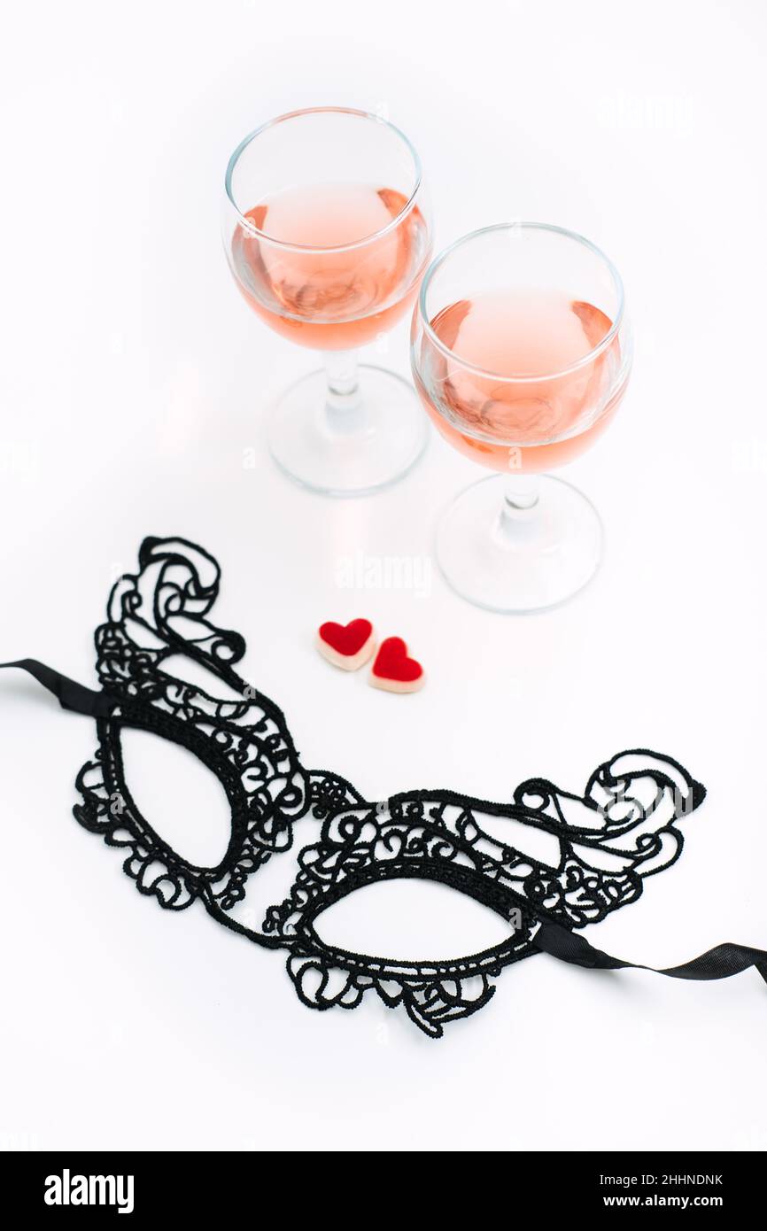 Table festive Saint-Valentin avec masque en dentelle noire, deux verres de vin rose et deux bonbons en forme de coeur rouge sur table blanche.Saint Valentin Banque D'Images