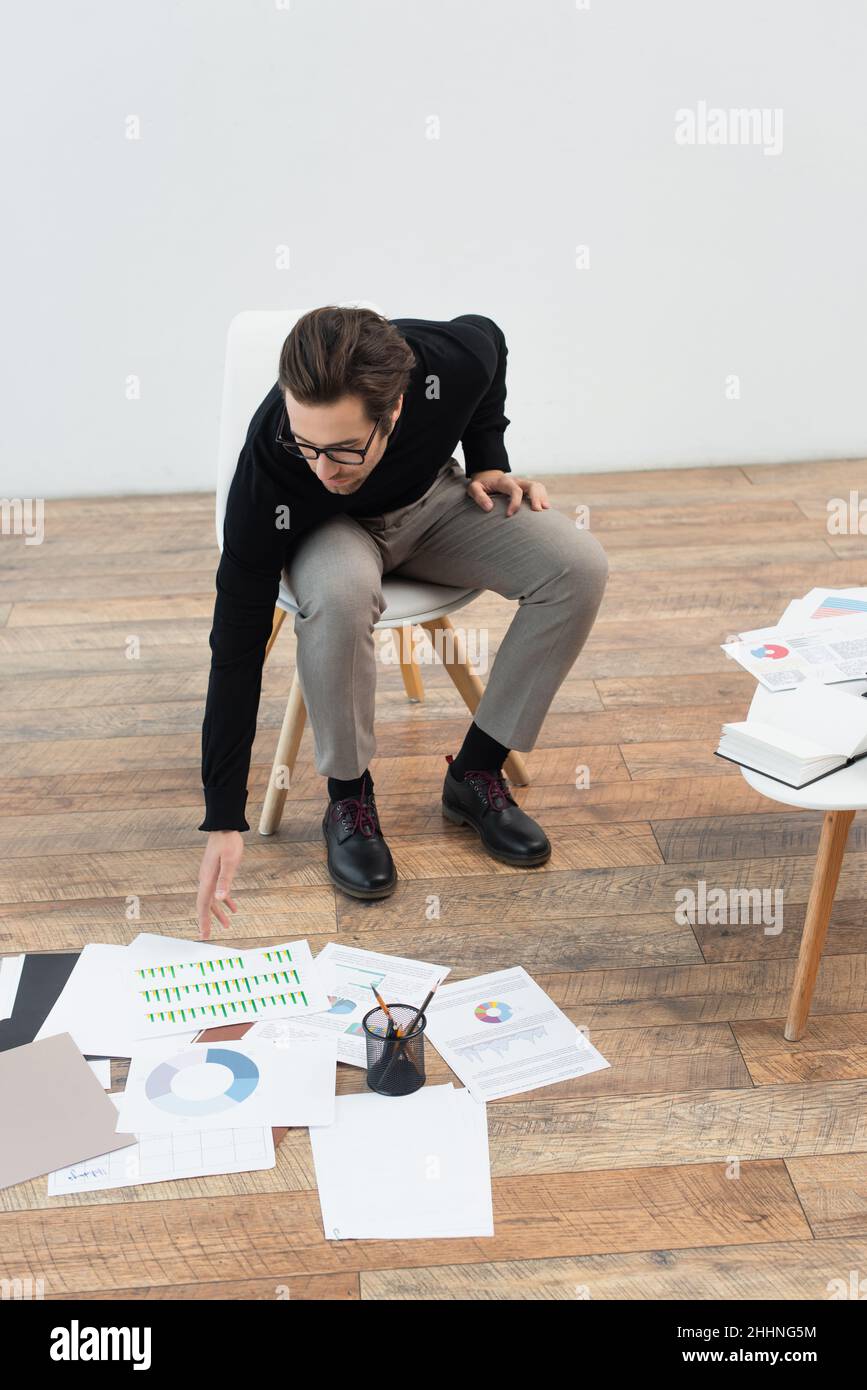 Homme Assis Sur Une Chaise Et Atteignant Des Documents Avec Des Infographies Sur Le Sol Photo 