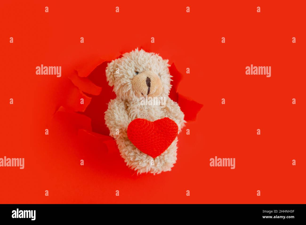Ours en peluche tenant le coeur rouge Hug dans les pattes.L'ours en peluche sort des trous de papier déchirés avec le symbole coeur d'amour pour la Saint-Valentin isolé sur le rouge Banque D'Images