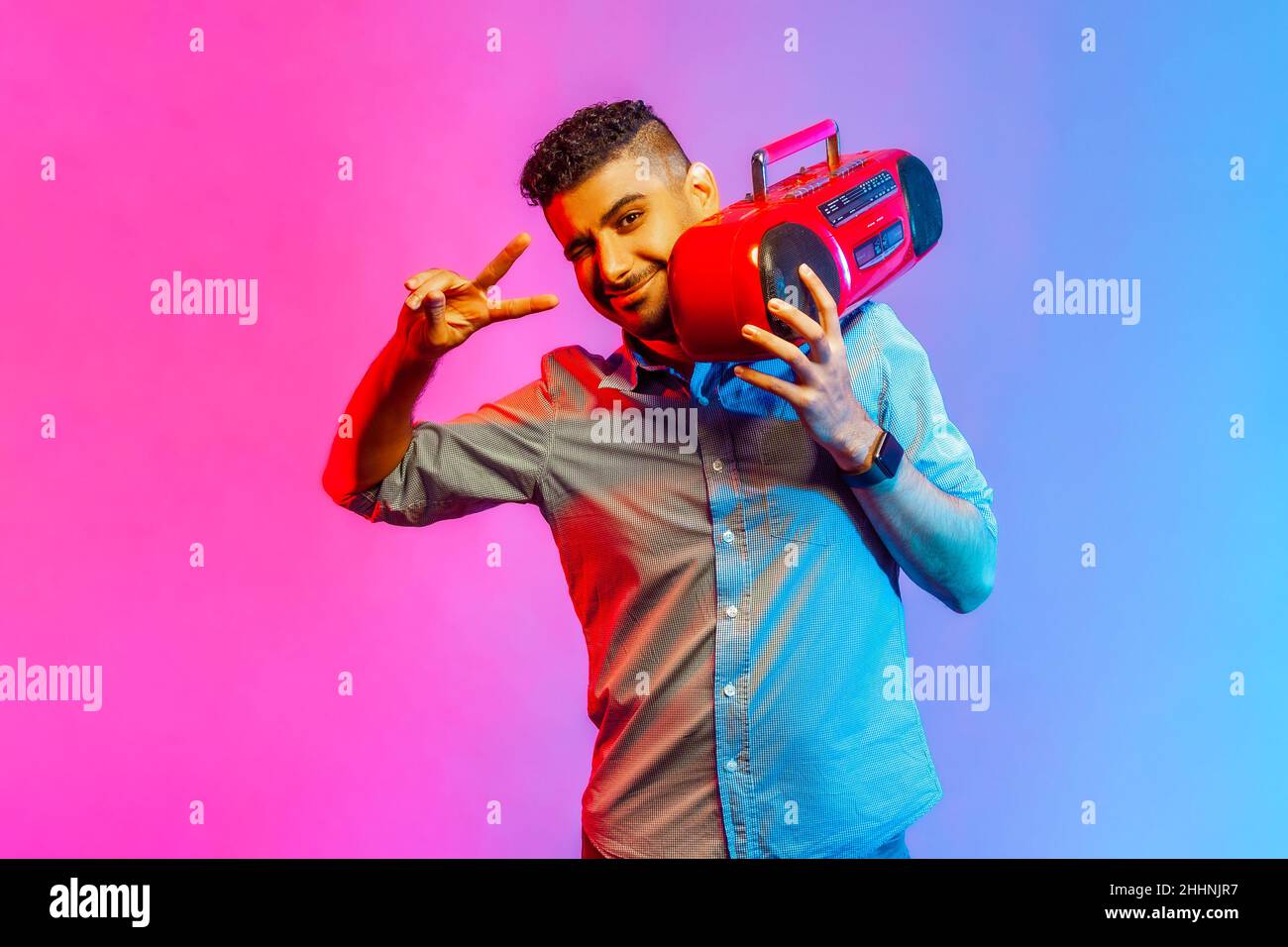 Portrait de l'homme heureux en chemise montrant le signe v avec les doigts tenant le magnétophone rouge sur l'épaule, en appréciant la musique préférée.Studio d'intérieur isolé sur fond de néon coloré. Banque D'Images