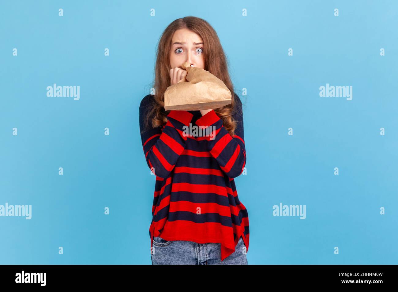 Malade femme portant un pull rayé style décontracté, respirant dans un sac en papier pour améliorer le bien-être, surmonter le stress, regardant l'appareil photo avec peur.Studio d'intérieur isolé sur fond bleu. Banque D'Images