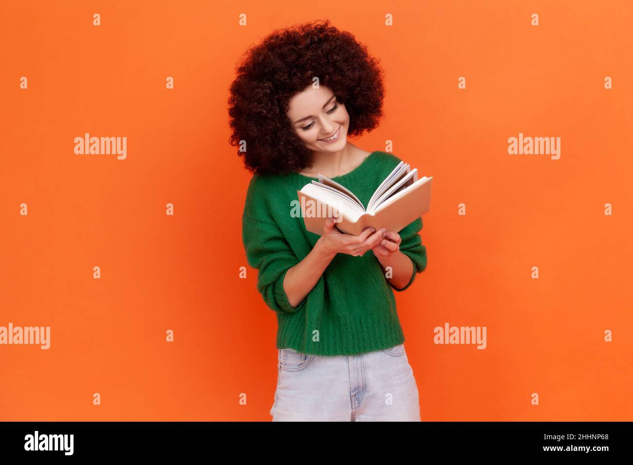 Bonne femme souriante avec la coiffure d'Afro portant vert décontracté style sweat livre de lecture avec intrigue intéressante, étudiant avec plaisir.Studio d'intérieur isolé sur fond orange. Banque D'Images