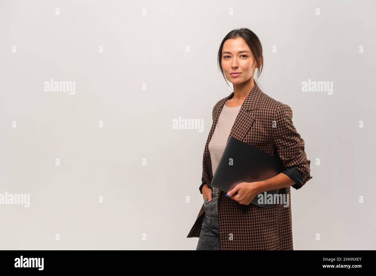 Femme asiatique élégante et confiante debout avec un ordinateur portable dans ses mains et regardant l'appareil photo sur fond gris.Portrait de produits et services de publicité sophistiqués pour femmes Banque D'Images