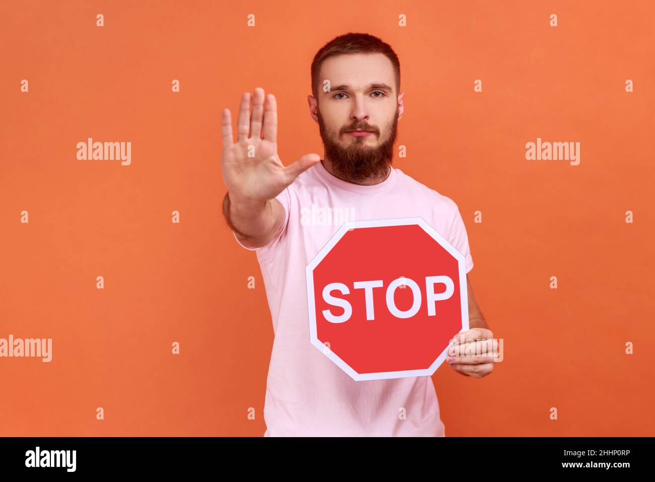 Portrait d'un homme à barbu et à la bosseux, montrant un geste d'arrêt et tenant un panneau rouge d'arrêt, interdictions et restrictions, portant un T-shirt rose.Studio d'intérieur isolé sur fond orange. Banque D'Images