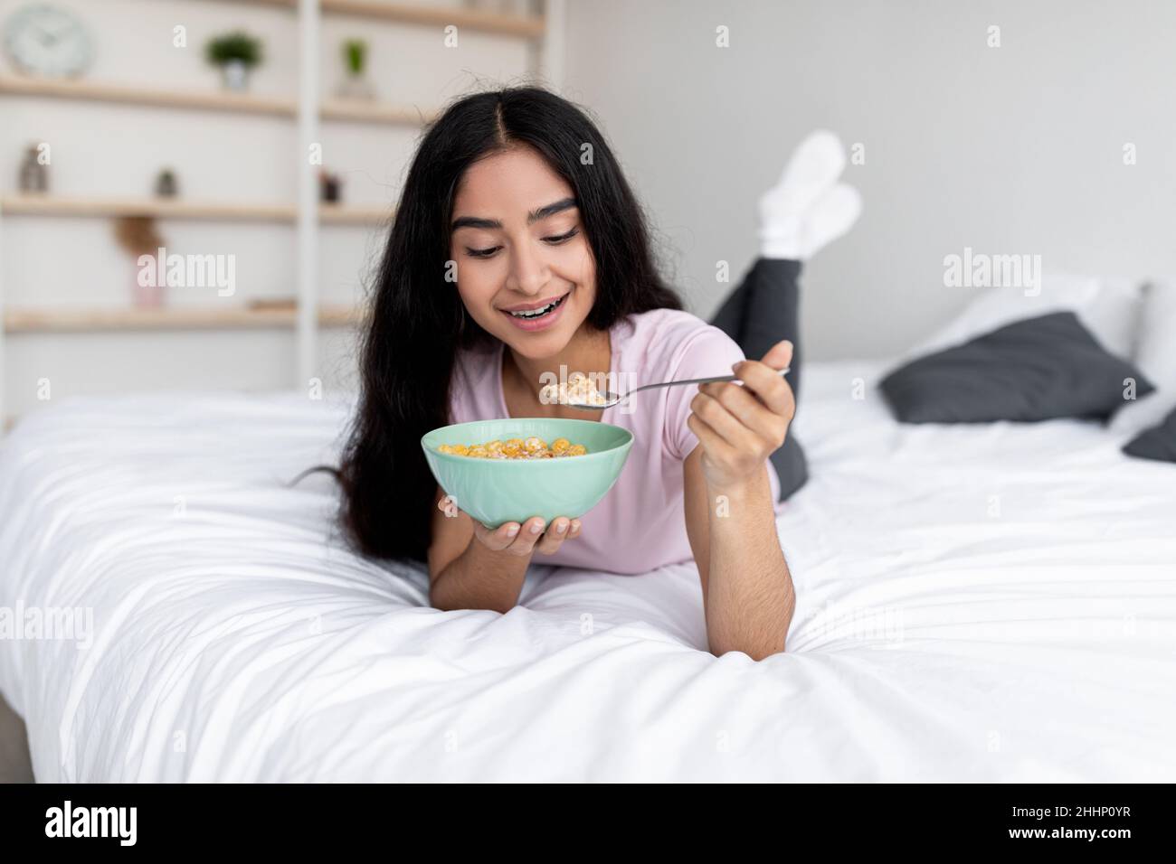 Portrait d'une jeune femme indienne mince mangeant des céréales avec du lait tout en étant allongé sur le lit à la maison, pleine longueur Banque D'Images