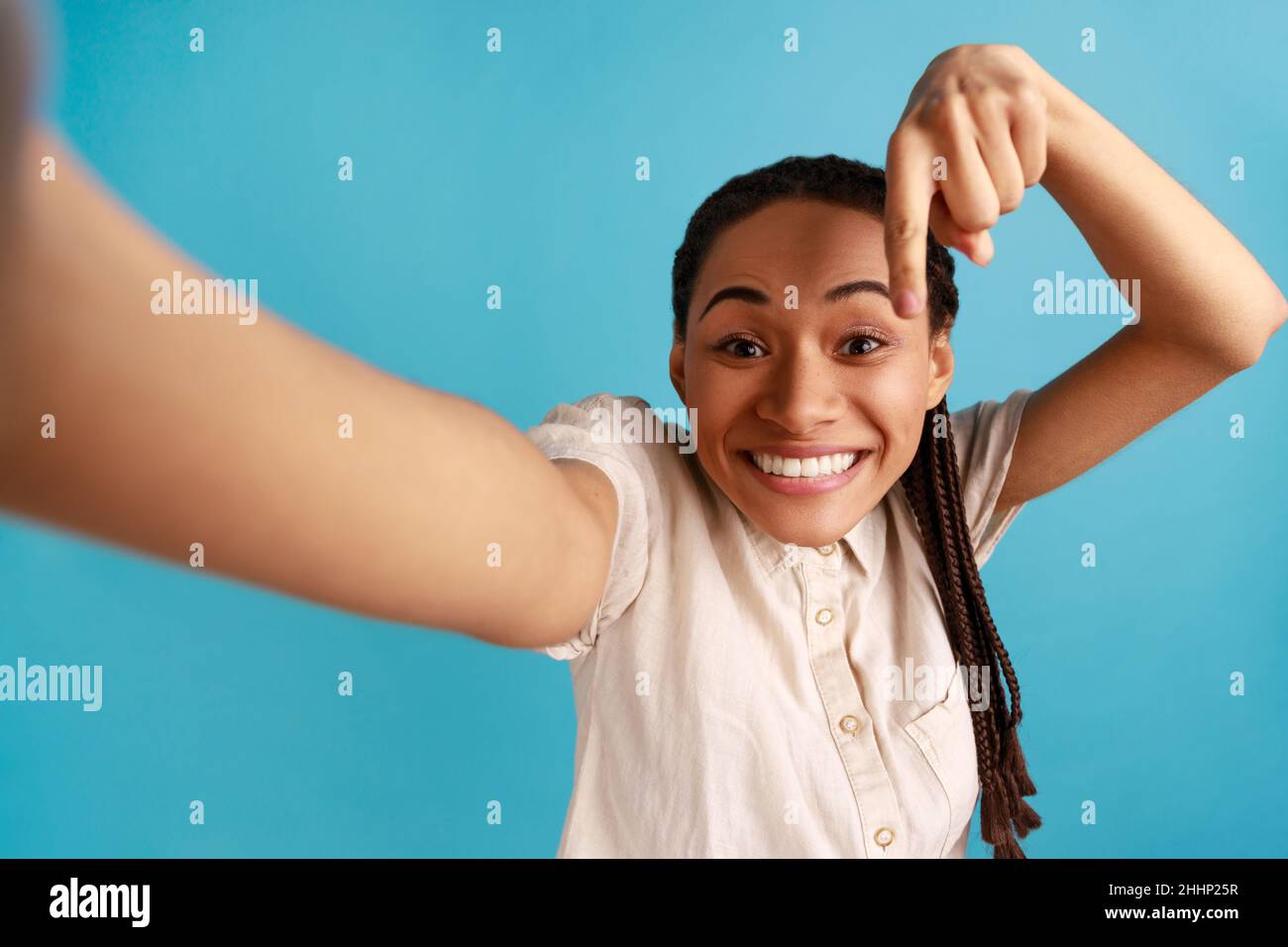 Portrait de femme souriante heureuse avec des dreadlocks faisant le selfie, pointant le doigt vers le bas, demandant de s'abonner, point de vue photo, portant une chemise blanche.Studio d'intérieur isolé sur fond bleu. Banque D'Images