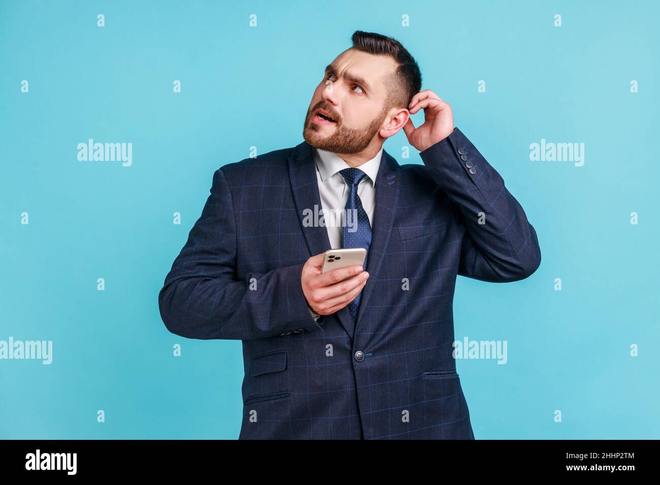 Homme d'affaires portant un costume de style officiel à la tête égratignant tenant le smartphone à la main, en envisageant la mise à jour de logiciels, en choisissant des tarifs appropriés.Studio d'intérieur isolé sur fond bleu. Banque D'Images