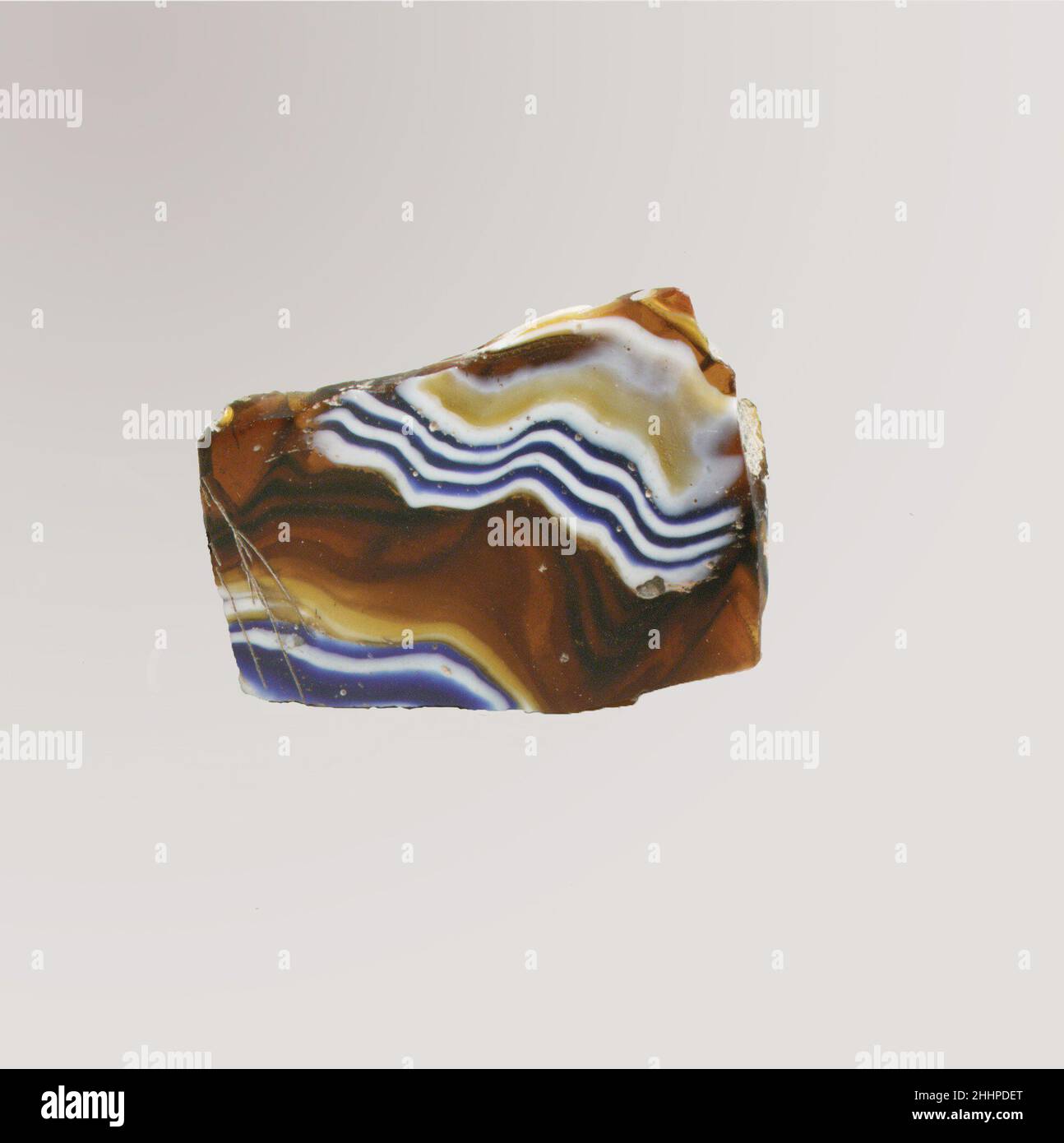 Fragment de bol nervuré en mosaïque de verre fin du 1st siècle C.-B.–début du 1st siècle A. D. fragment de corps romain.marron miel translucide, bleu cobalt et blanc opaque.cou légèrement disjoint; côté recourbé vers le bas.motif en mosaïque de ruban formé de sections d'une canne simple en sol brun avec fils ondulés blancs et bleus irréguliers en lignes parallèles;À l'extérieur, trois nervures verticales, assez espacées, avec des dessus aplatis et des bords extérieurs arrondis. Intérieur poli; piqûres de bulles et de fissures de surface à l'intérieur; morcellement et taches de mousse irisée crémeuse sur l'extérieur et les bords dentelés. Banque D'Images