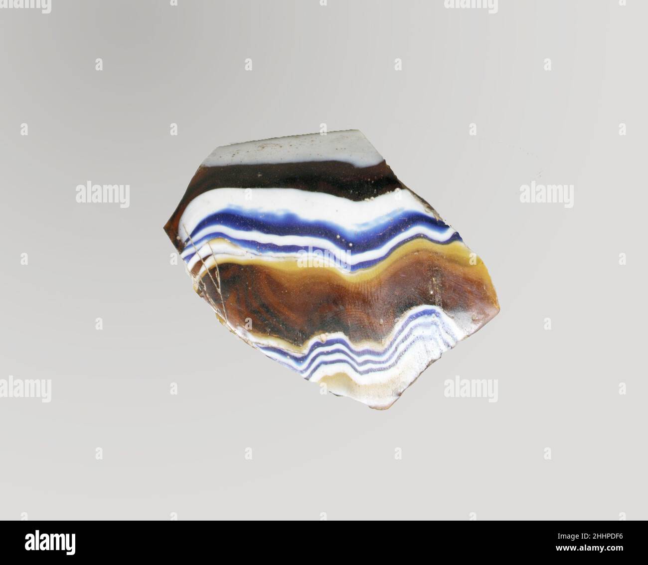 Fragment de bol nervuré en mosaïque de verre fin du 1st siècle C.-B.–début du 1st siècle A.D. fragment romain, probablement italien Rim.marron miel translucide, bleu cobalt et blanc opaque.rebord en saillie avec bord épais et arrondi;Côté droit en train de s'incliner vers le bas.motif en mosaïque de ruban formé de sections d'une canne simple au sol brun avec des fils irréguliers ondulés blancs et bleus dans des lignes horizontales parallèles; à l'extérieur, trois nervures verticales, espacées de manière inégale, avec des dessus aplatis et des bords extérieurs arrondis.intérieur poli; piqûres de bulles de surface et de fissures à l'intérieur;altération de la couche de mousse et d'irisée crémeuse o Banque D'Images