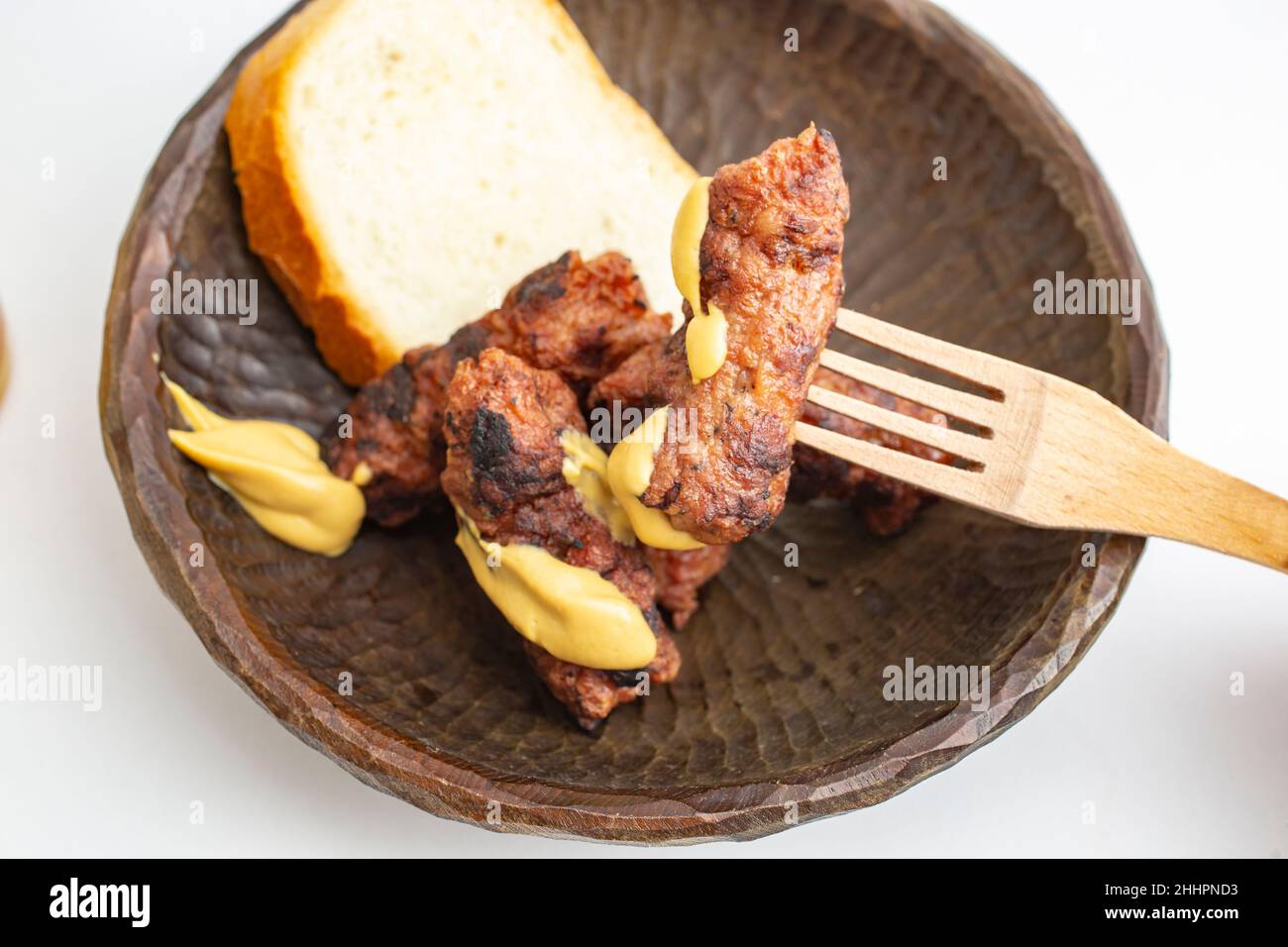 Roulés de viande hachés roumains, mici ou mititei sur un bol en bois sculpté avec de la moutarde et une fourchette traditionnelle en bois, isolée sur fond blanc Banque D'Images