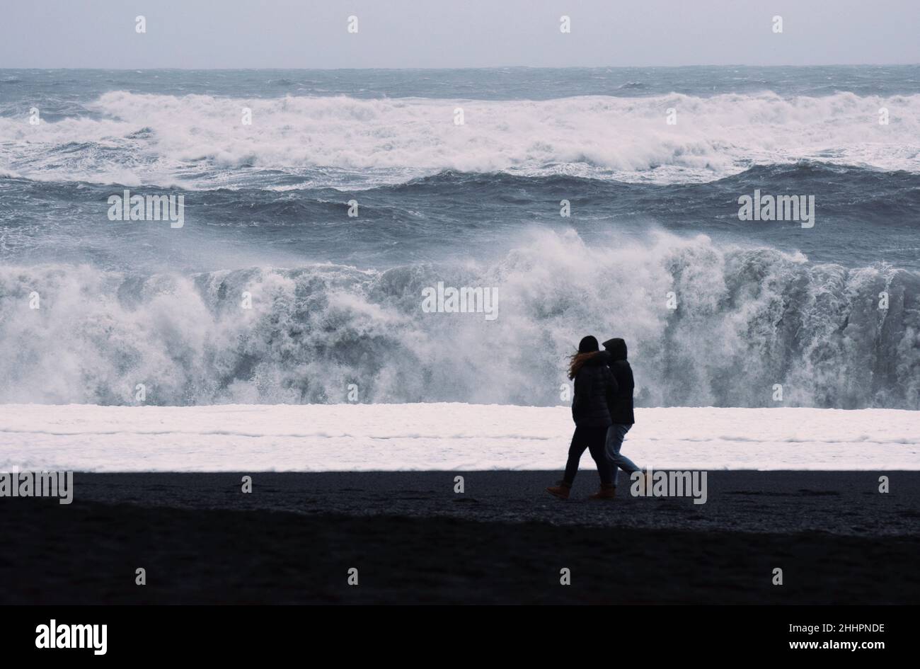 Deux touristes marchant sur la plage de sable noir de Reynisfjara près du petit village de pêcheurs de Vík dans le sud de l'Islande.Les vagues géantes éclipsent les marcheurs. Banque D'Images