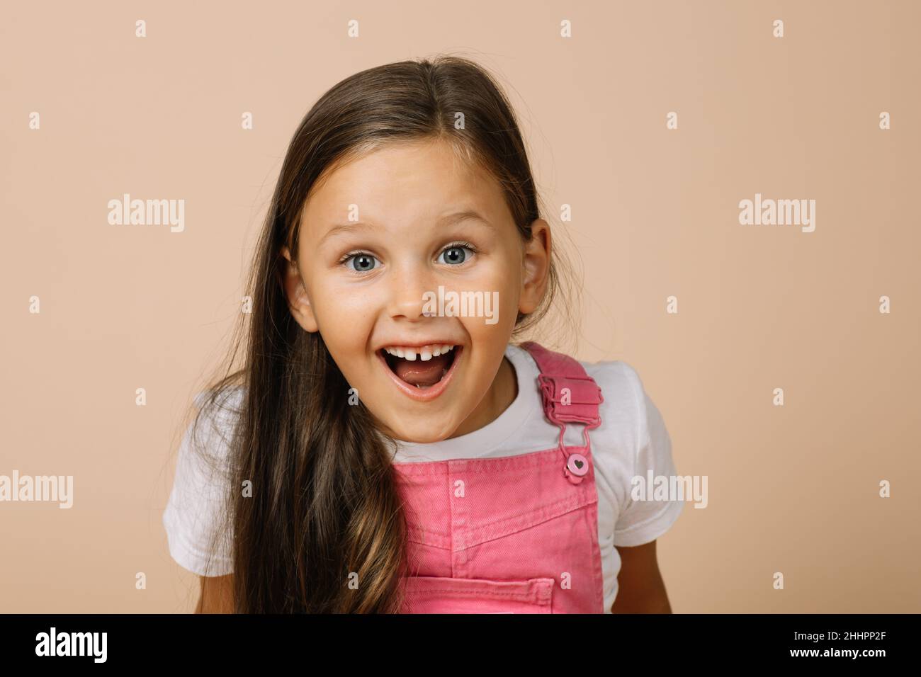 Petite fille avec des yeux surpris, bouche ouverte, sourire éclatant avec des dents et des sourcils relevés regardant l'appareil photo portant une combinaison rose vif et blanc Banque D'Images