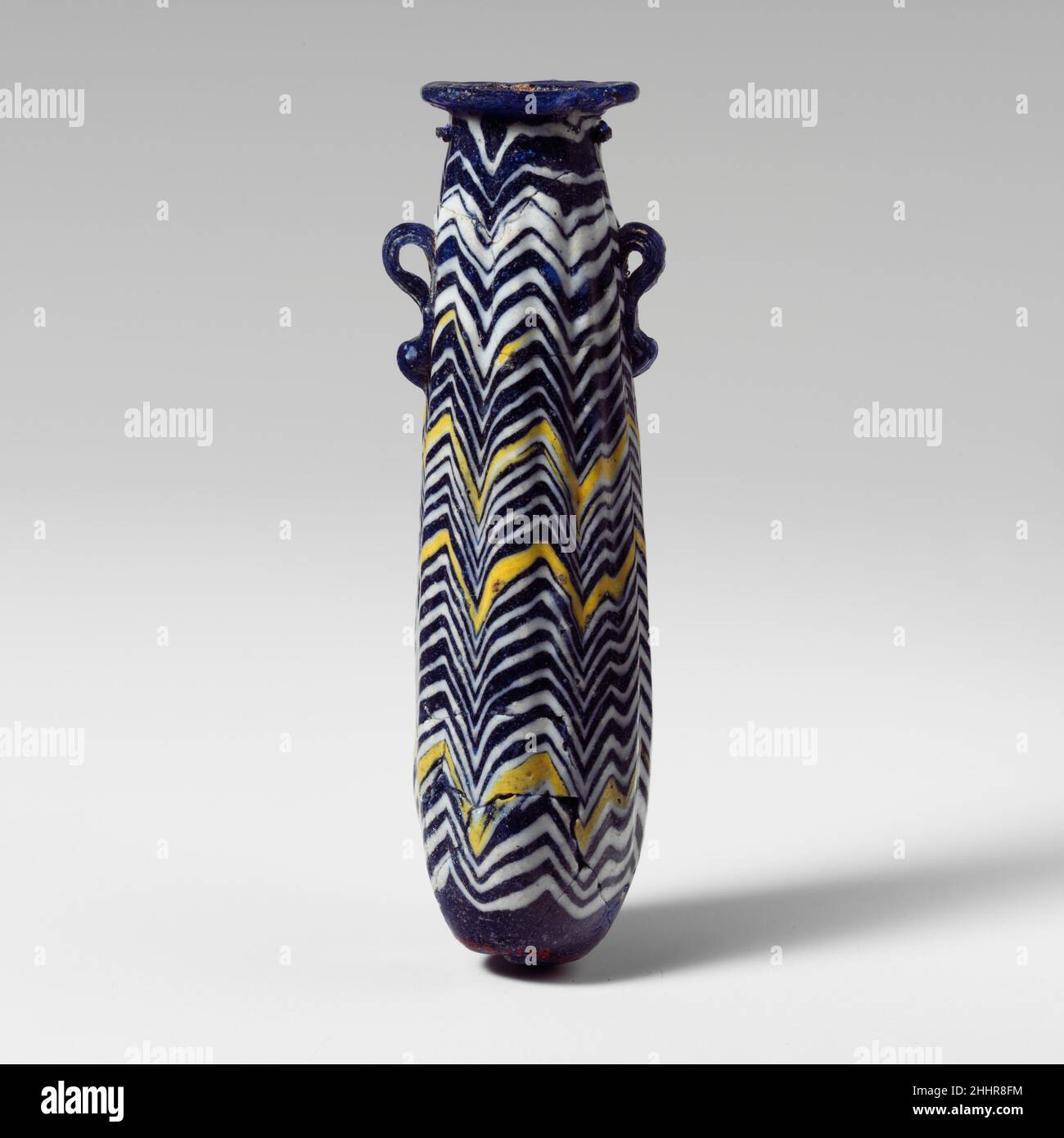 Verre alabastron (flacon de parfum) fin 6th–5th siècle C.-B. grec, Méditerranée orientale Bleu cobalt translucide, avec poignées de même couleur; traînées en blanc opaque et jaune opaque. Disque horizontal légèrement concave, formé comme une bobine, avec bord rugueux à la bouche; pas de cou ou d'épaule perceptible;Côté légèrement convexe sur corps cylindrique, en dépouille vers le haut ; fond arrondi, avec point central ; deux poignées circulaires verticales avec queues à boutons, appliquées sur la décoration de la piste. Traînée blanche fixée au cou sous le disque de la jante, enroulée de manière inégale, avec un motif zigzag étroit irrégulier, avec une rondelle profonde Banque D'Images