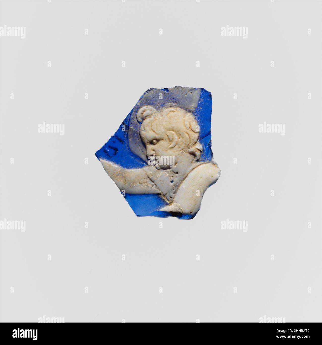 Fragment de caméo de verre fin du 1st siècle C.-B.–mi-1st siècle A.D. Roman bleu cobalt translucide avec revêtement blanc opaque.fragment de corps d'une tasse ou d'un bol relativement droit.en relief en blanc, tête d'une figure masculine jeune, peut-être Cupid, face à gauche mais regardant vers le bas,Avec le bras droit bien tendu avant lui et le bras gauche étendu vers le bas sur sa poitrine; un manteau fin, presque translucide, survole derrière sa tête.cassé de tous les côtés avec des fissures et des éclats à l'intérieur; mordillements, piqûres légères, et un léger vieillissement crémeux.marques de meulage rotatif à l'intérieur.Fragment de caméo en verre 249610 Banque D'Images