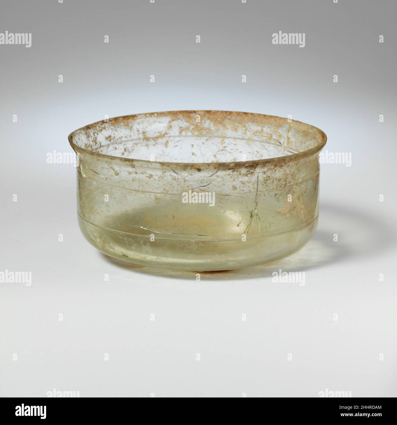 Bol en verre mi-1st–2nd Century A.D. Roman incolore avec du bleu vergeDébouchez, rebord inégal avec collet inférieur; côtés verticaux, arrondi à la base; fond avec base centrale surélevée avec un coup peu profond.Décoration de deux rainures parallèles de découpe de roue sur la carrosserie, espacées de 13 mm.une puce dans la jante et plusieurs fissures sur les côtés; peu de bulles;de légères taches de morcellement et d'intempéries, principalement à l'extérieur du fond; un peu d'enrobements de terre autour de la jante et sur les côtés à l'intérieur.Bol en verre 239875 Banque D'Images