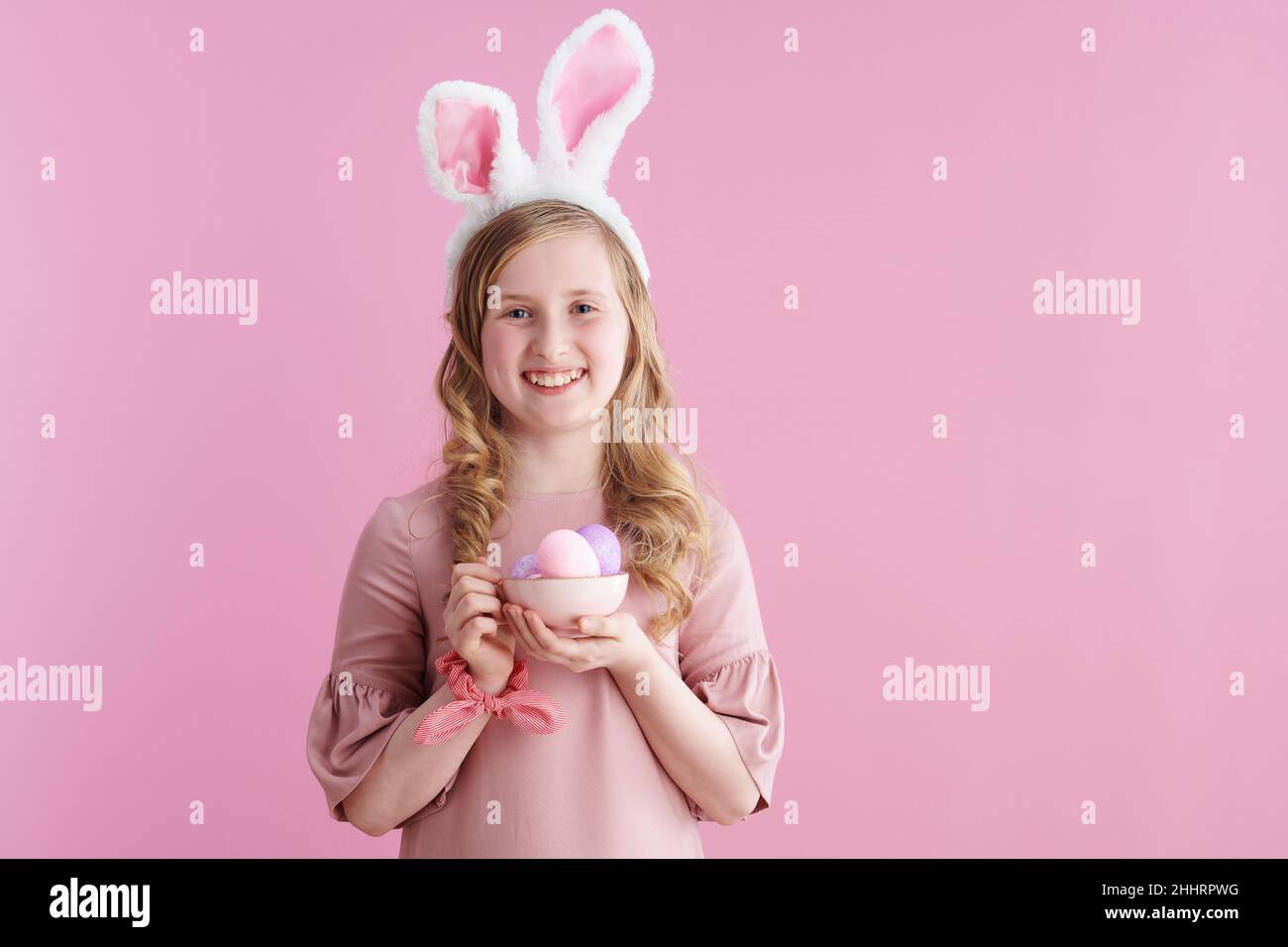 Portrait d'une jeune fille moderne souriante avec de longs cheveux blonds ondulés avec des oreilles de lapin et une tasse d'œufs de pâques isolés sur fond rose. Banque D'Images