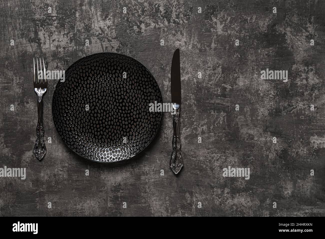 Définir la table.Assiette à dîner noire, couteau de table et fourchette sur surface texturée foncée.Vider la plaque en porcelaine près du couteau et de la fourchette vintage pour la conception alimentaire. Banque D'Images