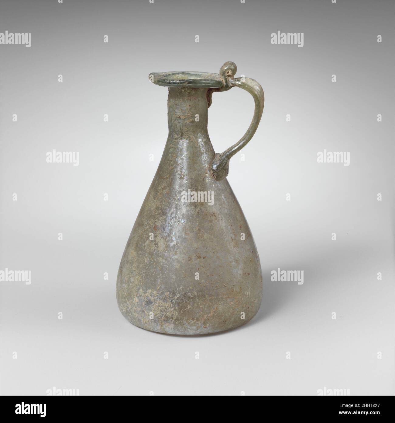 Verseuse en verre 1st–2nd Century A.D. Roman translucide bleu pâle vert, avec manche de même couleur.rebord tubulaire, replié, rond et intérieur, et pressé dans le haut de la bouche; col cylindrique court avec indentations irrégulières; côtés droits sur le corps conique; fond enfoncé;Poignée de sangle appliquée comme un coussin sur le dessus du corps, tirée dans une boucle et traîné sur le bord du bord et le haut du cou, avec une boucle verticale au-dessus.intact; quelques bulles de pinprick; morcellement, faible irience, limy enrouille et l'altération.Verseuse en verre 244658 Banque D'Images