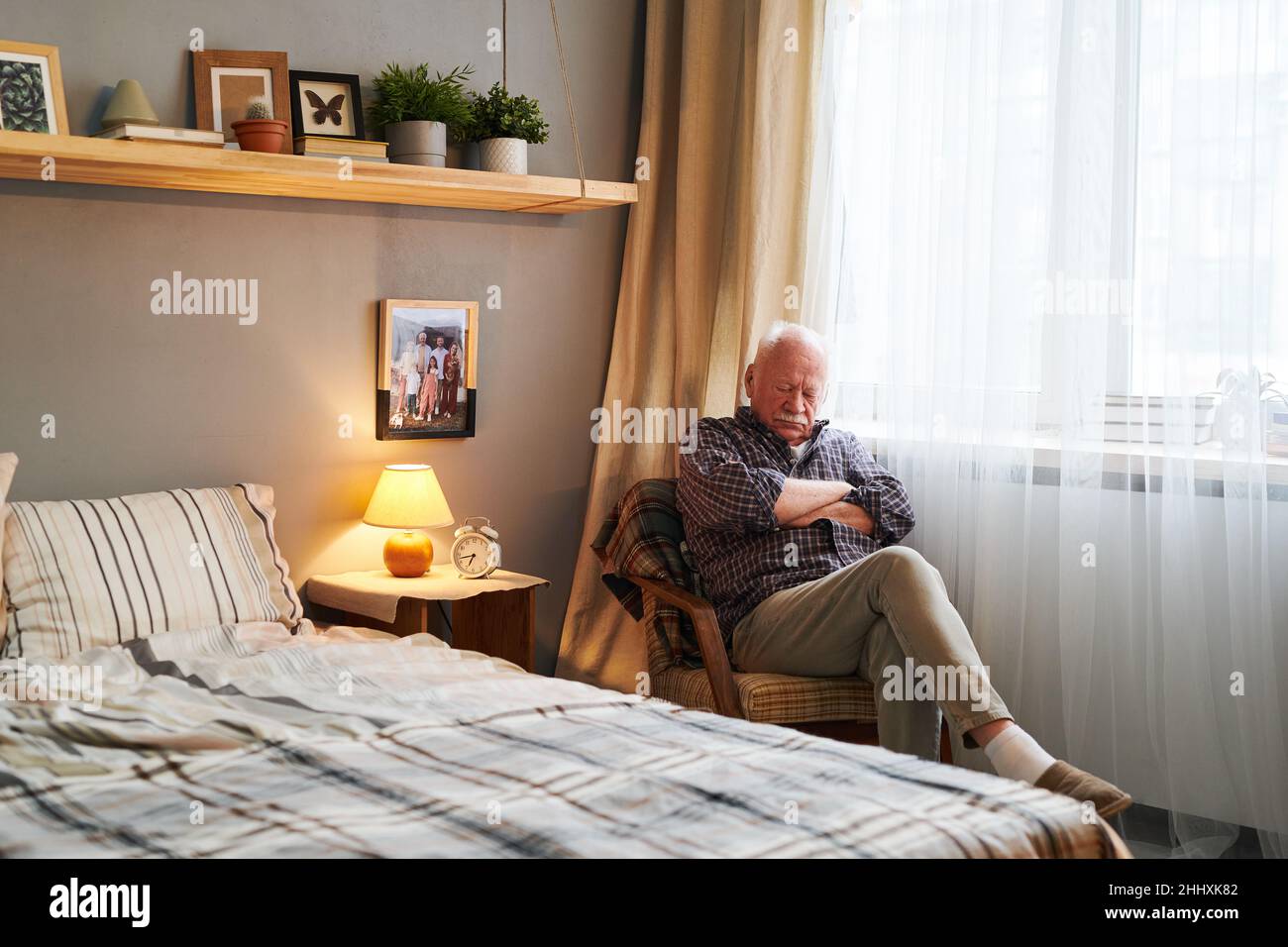 Homme âgé fatigué assis dans un fauteuil par la fenêtre et en train de passer la journée devant un lit double dans l'environnement de la maison Banque D'Images