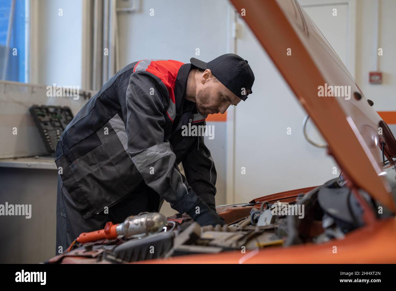 Jeune homme en tenue de travail se pliant sur le capot de voiture ouvert avec le moteur tout en le vérifiant avant du réparer dans l'atelier Banque D'Images