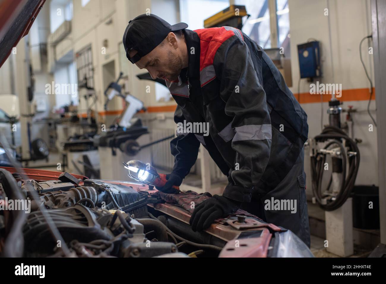 Moderne automécanique dans les vêtements de travail se pliant sur le capot de voiture ouvert tout en faisant le levage et la réparation du moteur dans le garage Banque D'Images