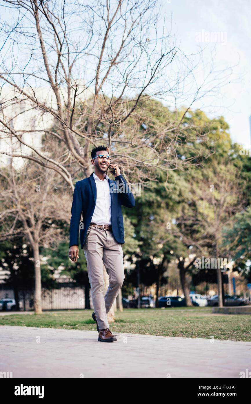 Grand cliché souriant homme noir entrepreneur marchant dans un parc tout en utilisant son téléphone faisant des affaires Banque D'Images