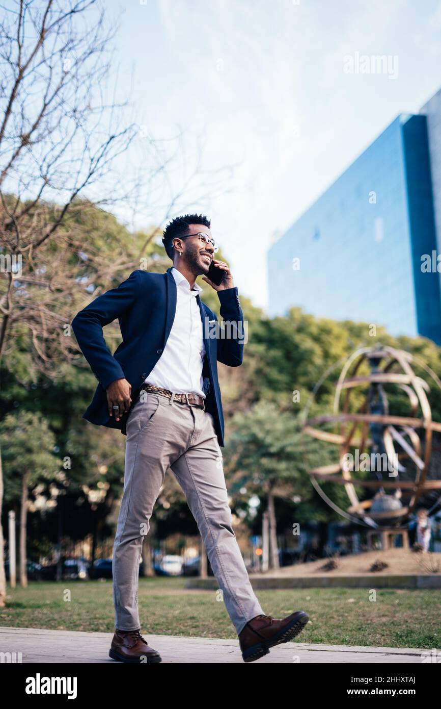 Homme d'affaires noir souriant et élégant se promenant dans un parc tout en utilisant son smartphone Banque D'Images