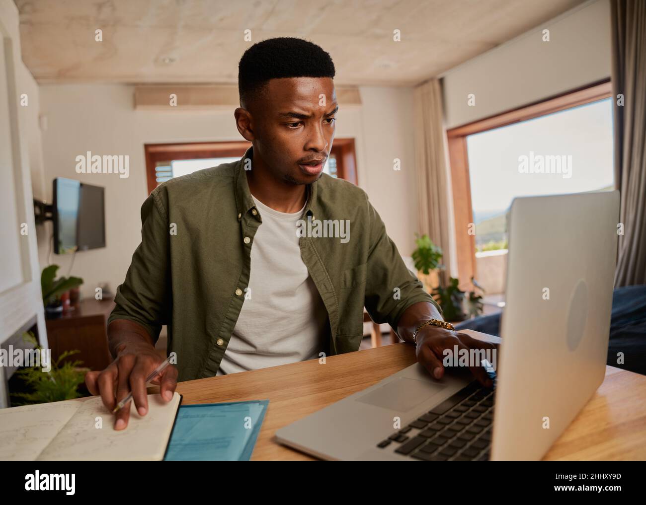 Jeune homme noir adulte stressé, prenant des notes à partir d'un ordinateur portable.Étudier à la maison, assis au comptoir de cuisine moderne. Banque D'Images