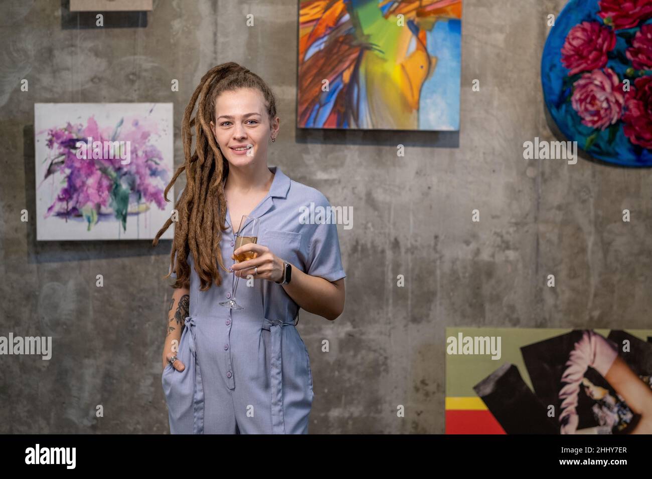 Portrait d'une jeune femme tenant une flûte de champagne et souriant à la caméra tout en se tenant en studio d'art avec des photos en arrière-plan Banque D'Images