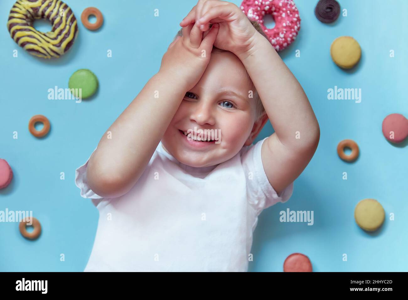 Mignon enfant souriant sur des beignets et des macarons français sur fond bleu.Concept de la Journée nationale du Donut.Concept d'enfance heureuse.Vue de dessus. Banque D'Images