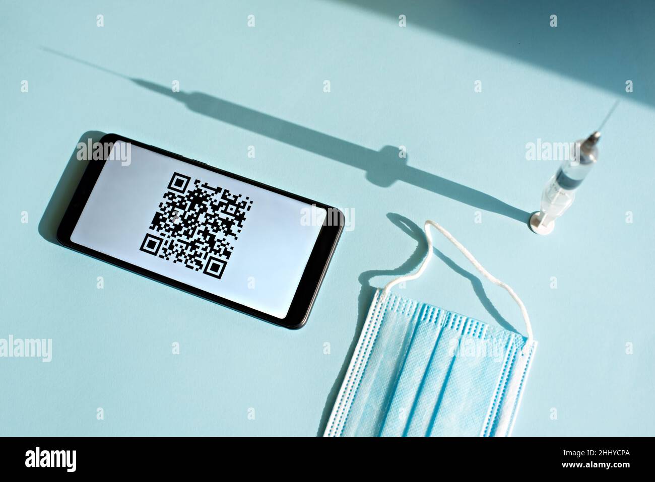 Smartphone avec code quar du certificat de vaccination sur l'écran, seringue et masque médical sur fond bleu clair. Banque D'Images