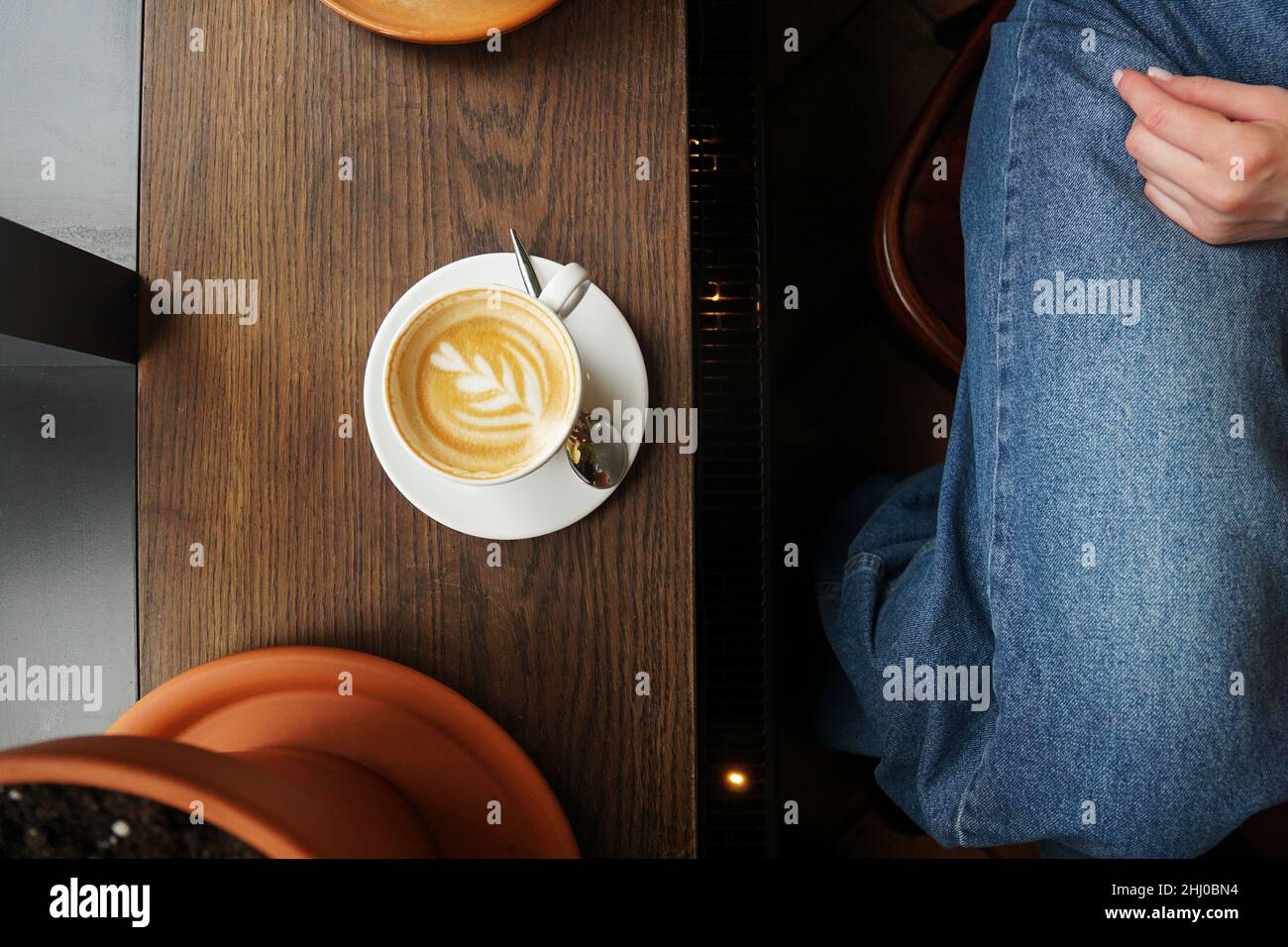 Service simple de cappuccino avec art floral en forme de cœur sur une table en bois sombre.Jean bleu d'une fille assise dans un café Banque D'Images