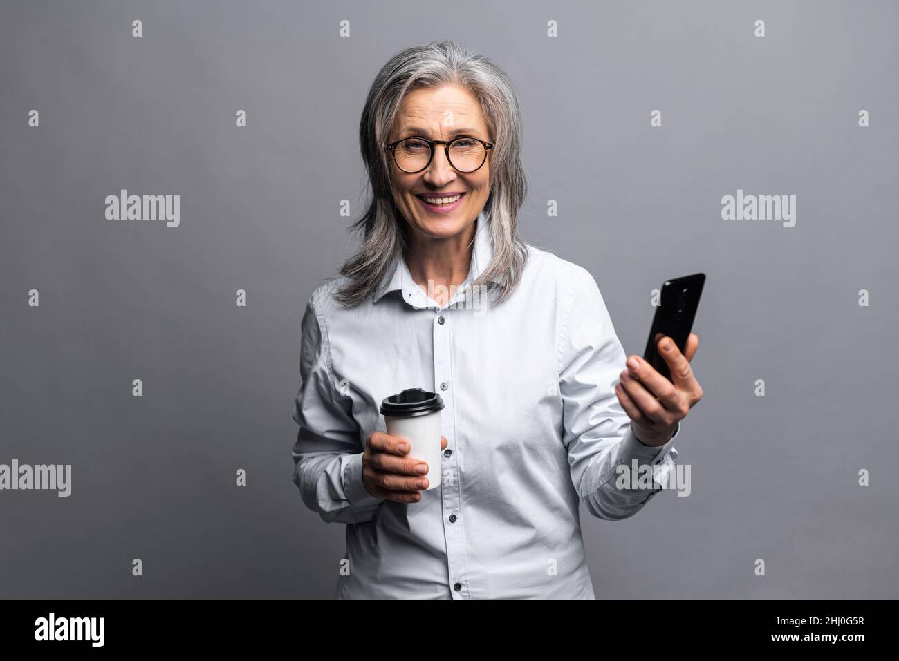 Charmante femme sénior en tenue formelle debout avec une tasse de papier de boisson chaude et smartphone isolé sur gris, se préparant à une bonne journée de travail.Une femme d'affaires mûre prend une pause-café Banque D'Images