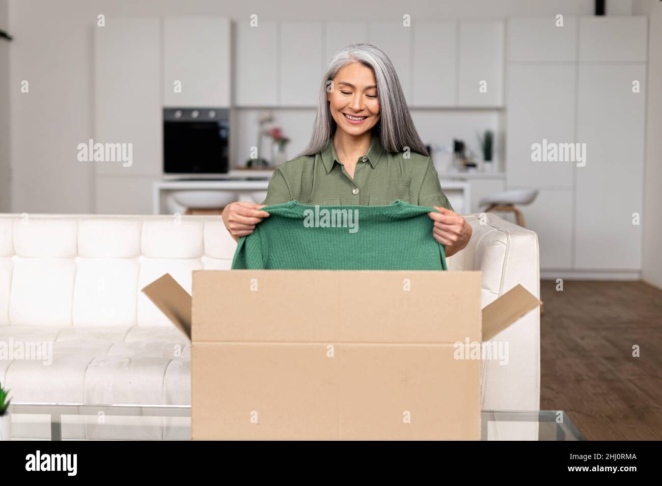 Positive mature femme asiatique debout avec boîte de carton et de prendre  de nouveaux vêtements, tout en se sentant curieux au sujet de l'article  commandé de la boutique en ligne.Client souriant satisfait
