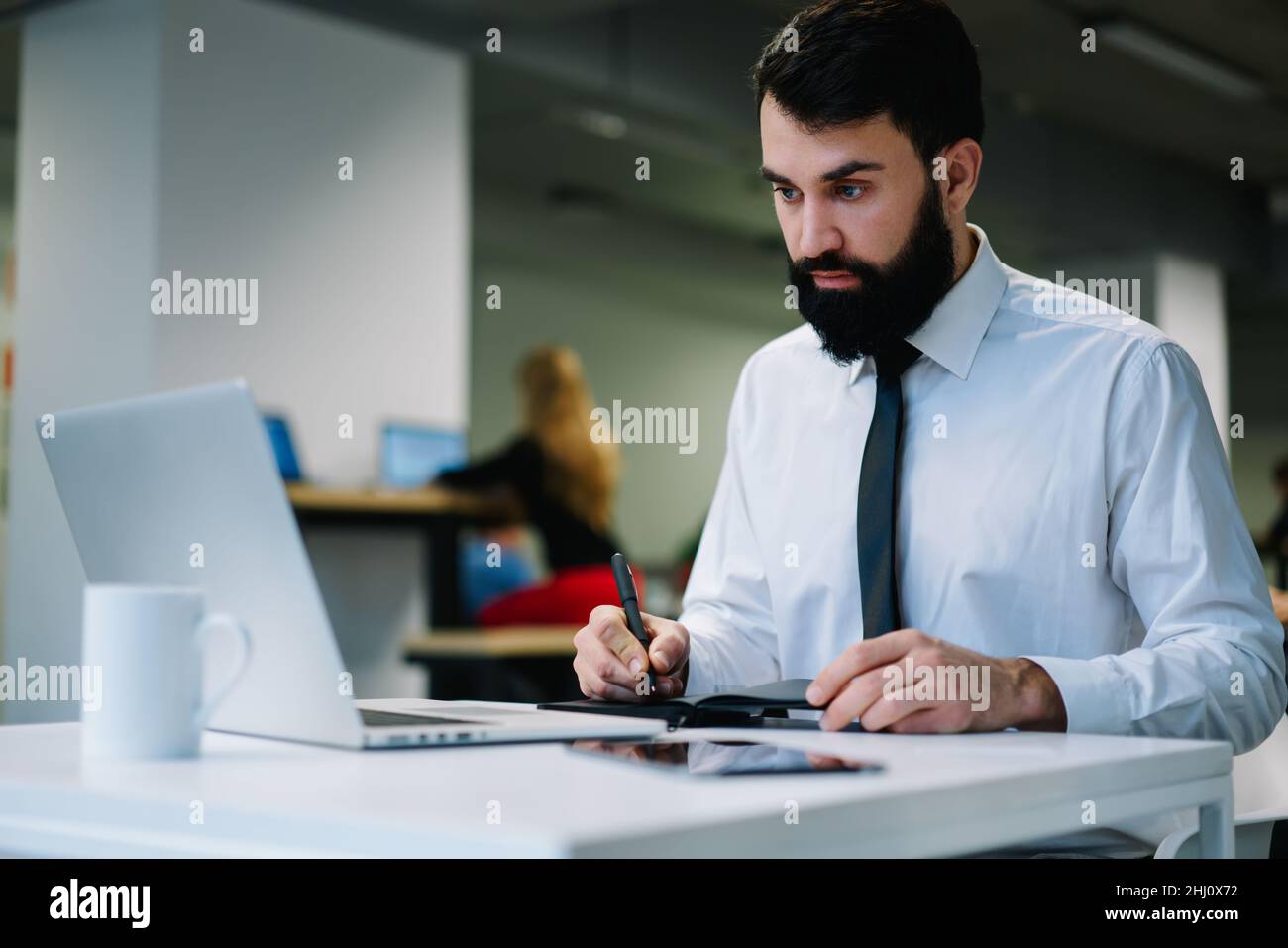 Homme d'affaires concentré prenant des notes sur un ordinateur portable tout en travaillant dans un espace de travail moderne Banque D'Images