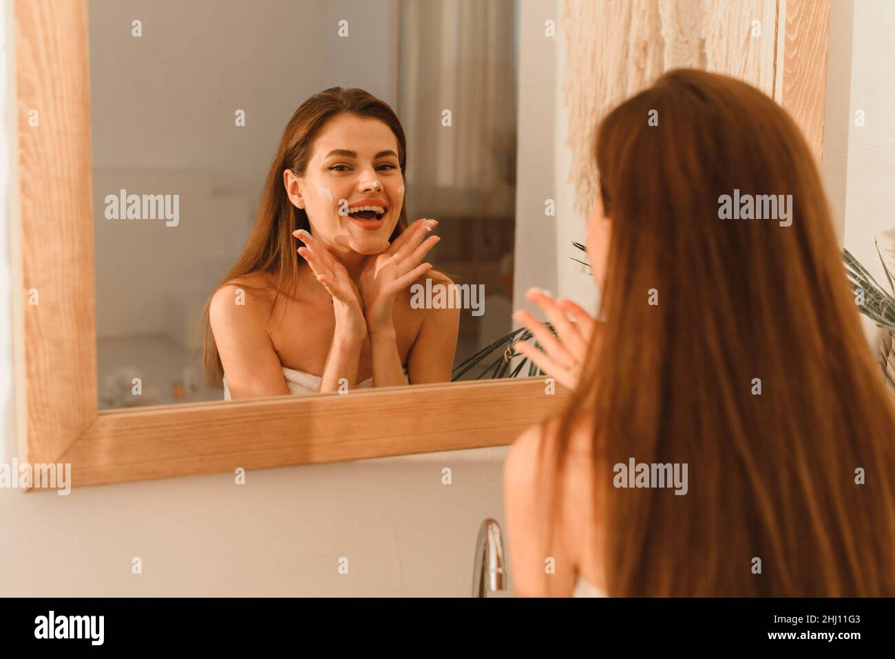 Rire jeune femme modèle appliquant hydratant à ses joues dans une salle de bains moderne Banque D'Images
